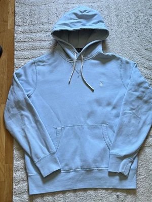 Ljusblå hoodie från Polo Ralph Lauren - Snygg ljusblå hoodie från Polo Ralph Lauren med klassisk huva och dragsko. Broderad logga på bröstet och stor känguruficka framtill. Mjuk bomullskvalitet som är skön att ha på sig. Perfekt för en avslappnad och stilren look.