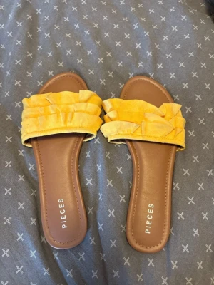 Gula sandaler med volang från Pieces - Snygga sandaler från Pieces i en härlig gul färg med volangdetaljer över foten. Sulan är brun och platt, och ovandelen är i mjukt tyg. Perfekta för en somrig look och lätta att matcha med olika outfits.