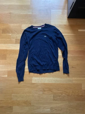 Mörkblå stickad tröja från Lacoste - Snygg mörkblå stickad tröja från Lacoste med rund halsringning och diskret logga på bröstet. Tröjan har långa ärmar och ribbade muddar vid ärmslut och nederkant. Perfekt till höst och vinter för en clean och klassisk look.