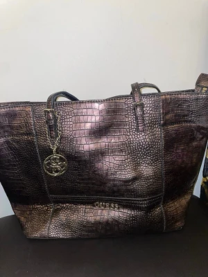Guess metallic  handväska - Säljer en stor handväska från Guess i metallic-brun färg med krokodilpräglat mönster. Väskan har dubbla axelband, en dekorativ Guess-logga i metall och dragkedjeficka på baksidan. Perfekt för dig som gillar statement-accessoarer.