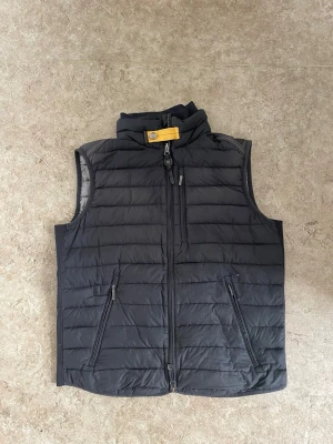 Svart dunväst från Parajumpers - Stilren svart dunväst från Parajumpers med klassisk gul rem och metallknapp vid kragen. Västen har tre dragkedjeförsedda fickor framtill och quiltad design. Perfekt för lager-på-lager och snygg till streetwear.