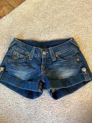 Blå jeansshorts från True Religion - Snygga blå jeansshorts från True Religion med uppvikta ben och dekorativa knappar på sidorna. Klassiska fickor fram och bak.