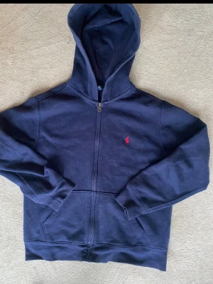 Mörkblå hoodie från Polo Ralph Lauren - Mörkblå hoodie med dragkedja från Polo Ralph Lauren. Klassisk design med huva, kängurufickor och röd broderad logga på bröstet. Tillverkad i mjukt bomullsmaterial som är skönt att ha på sig. Perfekt för en avslappnad och stilren look.