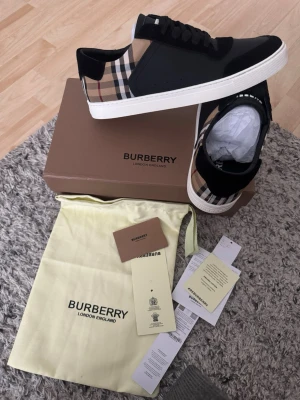 Burberry sneakers med rutigt mönster - Snygga sneakers från Burberry med klassiskt beige, svart och rött rutmönster på hälen, svart läder framtill och vita sulor. Skorna har svarta skosnören och låg profil. Perfekta för dig som vill ha en stilren och lyxig look. Storlek 44 passar 43 också och priset kan diskuteras. 