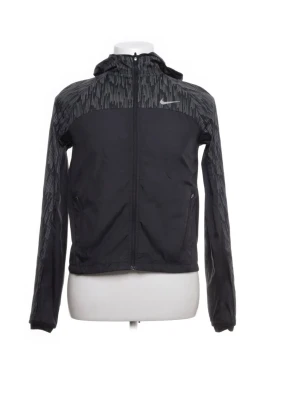 Svart vindjacka från Nike med mönster - Svart vindjacka från Nike med grått grafiskt mönster på axlar, huva och ärmar. Jackan har hel dragkedja, två fickor med dragkedja och en liten Nike-logga på bröstet. Perfekt för blåsiga dagar och sportiga outfits.