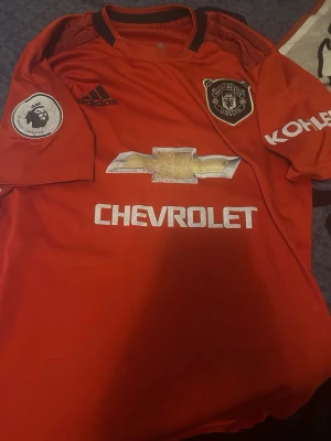 Manchester United matchtröja röd - Röd Manchester United matchtröja från Adidas med svarta detaljer. Tröjan har Premier League-märke på ärmen, klubbmärke på bröstet och sponsorloggor från Chevrolet och Kohler. Klassisk sportdesign i lätt och bekvämt material, perfekt för fotbollsträning eller support.