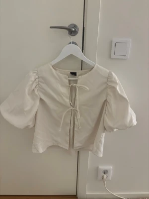 Vit blus med puffärm och knytning - Trendig vit blus med puffiga kortärmar och knytning framtill. Blusen har rund halsringning och är tillverkad i ett lätt, vävt material som ger en luftig känsla. Perfekt för en stilren och modern look.