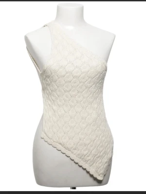 Vit stickad oneshoulder topp - Snygg vit stickad topp med oneshoulder-design och knytning i ryggen. Toppen har ett unikt mönster och asymmetrisk nederkant som ger en cool och trendig look. Perfekt för dig som vill sticka ut med en annorlunda topp.