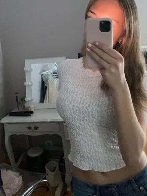 Vit croppad topp med volangkant - Söt vit croppad topp med volangkant och ribbad struktur. Toppen är ärmlös och har en lätt stretchig passform som sitter snyggt på kroppen. Perfekt att styla med jeans eller kjol för en trendig look.