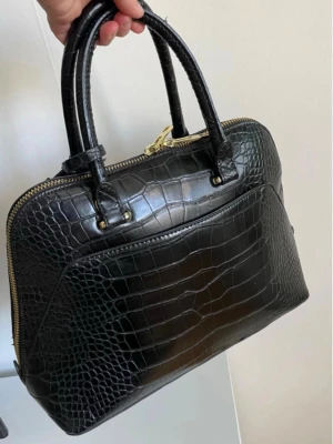 Svart stilren handväska Zara  - Stilren svart handväska med guldfärgade detaljer och dubbla handtag. Väskan har en praktisk ytterficka och dragkedja upptill. Perfekt accessoar för att lyfta din outfit.