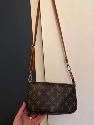  Monogram axelväska - Snygg axelväska i klassiskt brunt monogrammönster. Väskan har en avlång, rektangulär form och justerbar axelrem i läder. Tillverkad i canvas med detaljer i läder. 