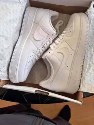 Nike Air Force 1 Low helvita sneakers - Klassiska Nike Air Force 1 Low i helvitt läder med ikonisk swoosh på sidorna. Skorna har perforerade detaljer vid tån, tjock vit sula och snörning framtill. Perfekta för en clean och stilren look.