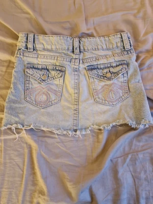 Ljusblå jeanskjol med fransig kant - Cool ljusblå jeanskjol med rå fransig nederkant och slitningar. Kjolen har två bakfickor med broderade detaljer och knappar, samt klassiska framfickor och bälteshällor. Materialet är denim och kjolen har en kort, rak passform.