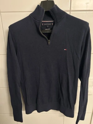 Mörkblå halvzip-tröja Tommy Hilfiger - Mörkblå stickad tröja från Tommy Hilfiger med halv dragkedja och hög krage. Liten broderad logga på bröstet och ribbade muddar. Tillverkad i mjuk premiumbomull för en skön känsla.