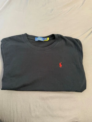 Svart t-shirt från Polo Ralph Lauren - Snygg svart t-shirt från Polo Ralph Lauren med klassisk rund hals och den ikoniska röda logotypen broderad på bröstet. Tillverkad i mjuk bomull som känns skön mot huden. Perfekt basplagg för en clean och stilren look.