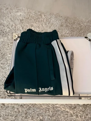 Gröna mjukisbyxor Palm Angels - Säljer ett par Palm Angels byxor i storlek L i en snygg grön färg.  Byxorna är i väldigt bra skick och har använts varsamt, utan några större slitningar eller skador i övrigt.  Det enda att nämna är att taggen baktill vid rumpan håller på att lossna, vilket syns på nära håll men påverkar inte funktionen eller helhetsintrycket vid användning.  Sitter riktigt bra och har den klassiska Palm Angels-stilen som ger en trendig och streetwear-inspirerad look. Perfekta för både vardag och mer stylade out