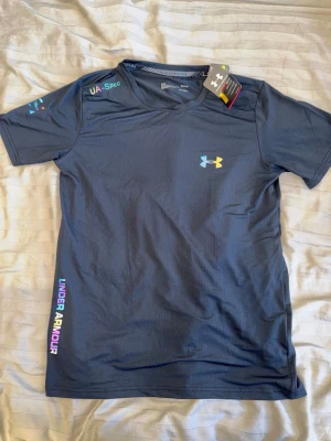 Svart Under Armour - Snygg svart t-shirt från Under Armour med färgglad logga på bröstet och text längs sidan. Tillverkad i syntetmaterial som andas, perfekt för träning. Kortärmad och med modern passform. Märkets logga syns även på ärmen. OBS STORLEK L