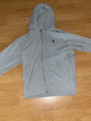 Grå hoodie från Polo Ralph Lauren - Snygg grå hoodie med dragkedja från Polo Ralph Lauren. Klassisk design med huva, två fickor framtill och den ikoniska lilla loggan broderad på bröstet. Perfekt för en avslappnad och stilren look.