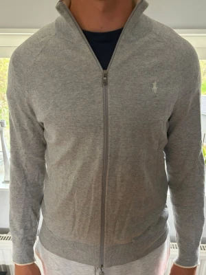Grå ziptröja från Ralph Lauren - Snygg grå ziptröja från Ralph Lauren med diskret logga på bröstet. Tröjan har hög krage, hel dragkedja och ribbade muddar. Perfekt för lager-på-lager och enkel att matcha med andra plagg. Storlek M och normal i strl.