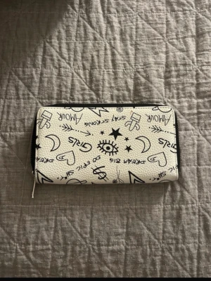 Vit plånbok med svart doodle-mönster - Cool vit plånbok med svart doodle-print med text som 'Girls', 'Dream Big', 'Stay Strong' och olika symboler som hjärtan, stjärnor och kaktusar. Plånboken har dragkedja runtom och är tillverkad i konstläder. Perfekt för dig som gillar unik och lekfull stil.