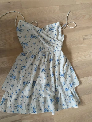 Blommig omlottklänning med volanger - Söt kortklänning med blått blommönster på vit botten. Klänningen har omlottdesign, smala axelband och volangdetaljer i kjolen. Perfekt för sommarens alla tillfällen. Resår i ryggen för skön passform.