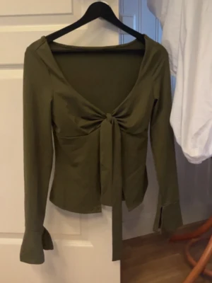 Olivgrön långärmad topp med knyt - Trendig olivgrön topp med långa ärmar och snygg knytning framtill. Djup v-ringning och lätt croppad passform ger en modern look. Perfekt att styla med både jeans och kjol för en cool vibe. Aldrig använd 