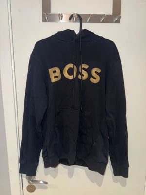 Svart hoodie med guldtext BOSS - Svart hoodie från BOSS med stor guldfärgad text på bröstet. Insidan av huvan har guldfärgad detalj. Perfekt för en chill och stilren look.