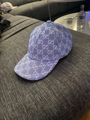 Blå Gucci keps med GG-mönster - Säljer en blå keps från Gucci med det klassiska vita GG-monogrammet över hela kepsen. Kepsen har böjd skärm och justerbart band bak. Materialet är denimliknande tyg med vit insida och tydliga Gucci-taggar. Perfekt för dig som vill ha en stilren och lyxig accessoar.