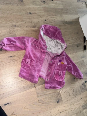 Rosa fleecejacka med huva Baby Club - Söt rosa fleecejacka från Baby Club med huva och tryckknappar. Jackan har två fickor med rosettdetaljer och små broderade figurer på bröstet. Perfekt för kyliga dagar och enkel att ta på.