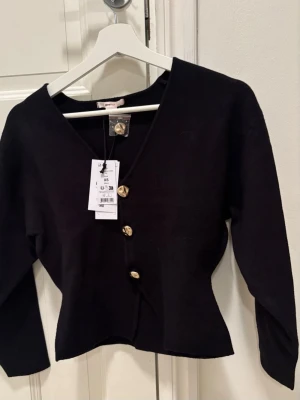 Gina Tricot 14+ Svart kofta med guldknappar - Snygg svart kofta från Gina Tricot, Sculpture Knitted Top, med djup V-ringning och fyra dekorativa guldknappar framtill. Modellen är figurnära med långa ärmar och mjukt material som ger en stilren look. Perfekt att matcha med jeans eller kjol. Ny med prislapp kvar. Slutsåld på Gina Tricot. 