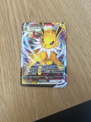 Jolteon VMAX Pokémonkort - Säljer ett färgstarkt Jolteon VMAX samlarkort från Pokémon. Kortet har en glansig yta med motiv av Jolteon i gult och vitt, samt detaljer i blått, rött och silver. Texten är på spanska och kortet har 300 HP. Perfekt för samlare eller Pokémonfans!