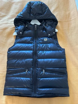 Svart dunväst med huva från Bvaldi - Snygg svart dunväst från Bvaldi med avtagbar huva och två dragkedjefickor framtill. Västen har quiltad design, hög krage och en logga på bröstet. Perfekt för lager på lager och håller dig varm under kyliga dagar.