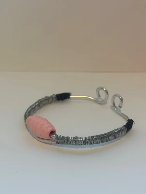 Silverfärgad armband med rosa detalj - Armband gjord av silverfärgad metalltråd med en rosa räfflad pärla. Armbandet är gjord av silverfärgad metalltråd som är böjd i ändarna. Runt metalltråden är en mindre metalltråd virad runt samt en svart metalltråd nära ändarna. 
