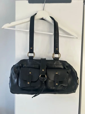 Svart handväska med fickor - Stilren svart handväska i läderimitation med två framfickor och guldfärgade detaljer. Väskan har dubbla axelband och stängs med rem och spänne. Invändigt finns ett stort fack och en innerficka med dragkedja. Perfekt för dig som gillar praktiska detaljer och klassisk look. Aldrig använt 