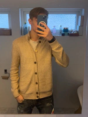 Beige kofta från massimo dutti - Säljer en beige stickad kofta med grovstickade axlar och bruna knappar framtill. Koftan har ståkrage och ribbade muddar vid ärmslut och nederkant. Perfekt för lager på lager och enkel att matcha med olika outfits.