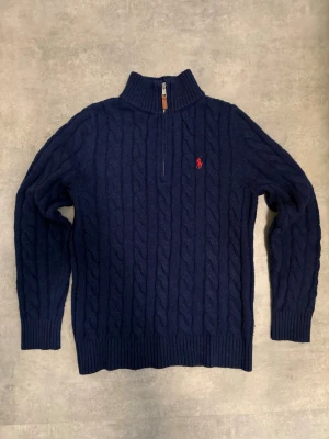 Mörkblå kabelstickad tröja Polo Ralph Lauren - Mörkblå kabelstickad tröja från Polo Ralph Lauren med halv dragkedja och hög krage. Tröjan har ett rött broderat logotyp på bröstet och ribbade muddar vid ärmslut och nederkant. Perfekt för lager på lager och riktigt snygg till jeans.