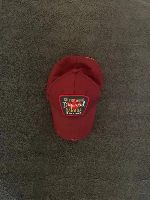 Vinröd keps från Dsquared2 - Säljer en vinröd keps från Dsquared2 med broderad patch framtill där det står 'City of Wood Dsquared Canada'. Kepsen har klassisk böjd skärm och justerbar rem bak. Tillverkad i bomull och har detaljer i gult och vitt på framsidan.