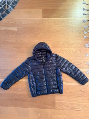 Svart dunjacka med huva från Replay - Snygg svart dunjacka från Replay med glansig finish och fast huva. Jackan har två sidofickor med dragkedja och quiltad design som ger en modern look. Perfekt för kalla vinterdagar och enkel att matcha med olika outfits.