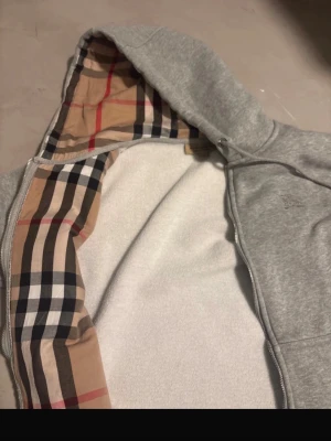 Grå hoodie från Burberry med dragkedja - Säljer en grå hoodie från Burberry med klassiskt rutigt foder i huvan. Hoodien har dragkedja framtill, snörning vid huvan och mjukt vitt innerfoder. Perfekt för dig som gillar exklusiva märken och stilrena detaljer.