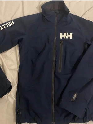Mörkblå Helly Hansen vindjacka - Snygg mörkblå vindjacka från Helly Hansen med vit HH-logga på bröstet och HELLY på ärmen. Använd max 1 gång säljer då den är för liten för mig.