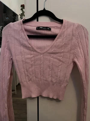 Ljusrosa stickad croptröja XS - Söt ljusrosa stickad croptröja från New Yorker med V-ringning och långa ärmar. Tröjan har ett ribbstickat midjeband och ett diskret kabelstickat mönster. Perfekt för dig som gillar en feminin och trendig stil.