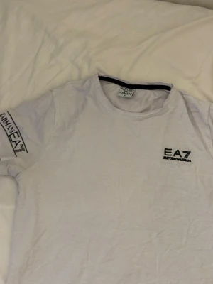 Vit EA7 t-shirt från Armani - Säljer en vit t-shirt från EA7 Emporio Armani med tryck på bröstet och båda ärmarna. T-shirten har rund hals och korta ärmar. Perfekt för dig som gillar stilrena och klassiska plagg med tydlig logga.