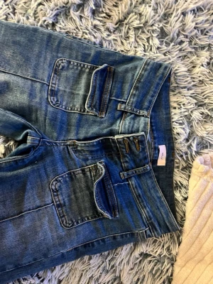 Blå jeans med stora fickor från Nelly - Säljer ett par blå bootcut jeans från Nelly med stora, fyrkantiga fickor framtill. Jeansen har dubbla knappar i midjan och markerade sömmar längs benen. Perfekta för en avslappnad och trendig stil. Trasiga nere vid fötterna, därav billigare pris. 