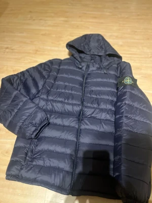 Mörkblå pufferjacka från Stone Island - Säljer en mörkblå pufferjacka från Stone Island med huva och dragkedja. Jackan har det klassiska Stone Island-märket på ärmen och är quiltad för extra värme. Perfekt för dig som vill ha en snygg och funktionell jacka med streetkänsla.