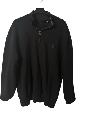 Svart halvzip tröja Polo Ralph Lauren - Säljer en svart stickad tröja från Polo Ralph Lauren med halv dragkedja och diskret logga broderad på bröstet. Tröjan har ribbade muddar och krage med vit rand. Perfekt för dig som gillar klassisk och stilren design.