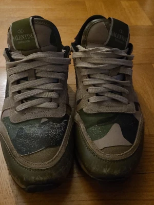 Valentino camo sneakers - Snygga sneakers från Valentino med kamouflagemönster i grönt, beige och svart. Skorna har beige snörning, detaljer i mocka och läder samt nitar på hälen. Perfekt för dig som vill ha en unik och trendig look.