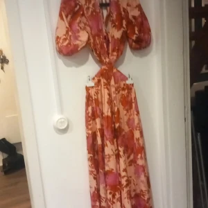 Blommig långklänning med puffärm - Säljer en långklänning med blommigt mönster i rosa och rött. Klänningen har puffärmar, v-ringning och markerad midja med cut-out-detalj. Perfekt för dig som vill sticka ut med en färgstark och unik look.