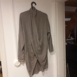 Grå lång kofta med draperad front - Säljer en grå lång kofta med draperad front och hög krage. Koftan har långa ärmar och ett mjukt, tunt material som faller snyggt. Perfekt för lager på lager och en avslappnad stil.