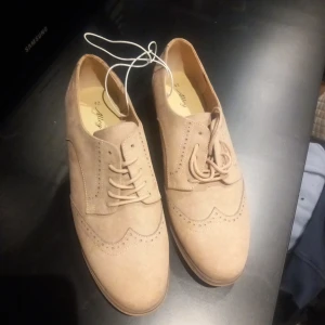 Beige mockaloafers med broguemönster - Säljer ett par snygga beige lågklackade i mocka med klassiskt broguemönster och rundad tå. Skorna har snörning och låg klack, perfekta för dig som gillar stilrena detaljer. Storlek 42 står i skon. Passar till många olika outfits.