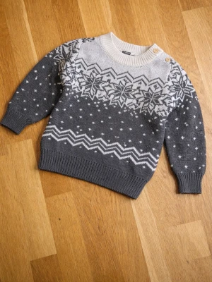 Grå stickad tröja med snöflingor - Barn tröja Mysig grå stickad tröja med vitt snöflingemönster och zigzag-detaljer. Tröjan har rund halsringning med ribbad kant och två knappar på axeln. Långa ärmar och ribbade muddar vid ärmslut och nederkant. Perfekt för kalla dagar.